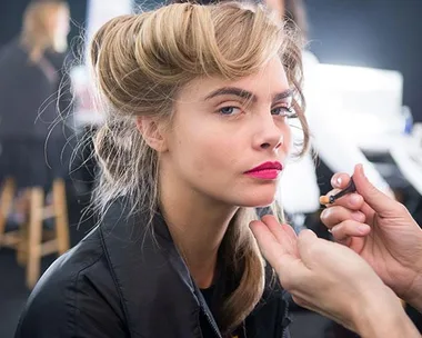 Model with styled hair and bright lipstick applies makeup backstage.