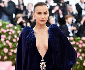 A woman in a deep blue gown poses on a floral-themed red carpet with photographers in the background.