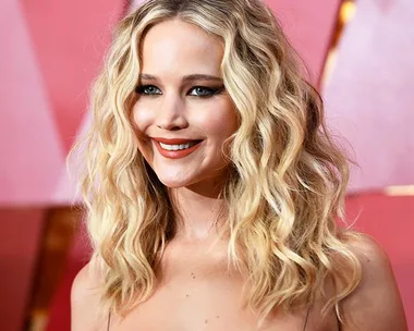 Actress with wavy blonde hair smiling on a red carpet background.