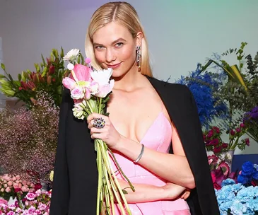 A woman in a pink dress and black jacket holds a bouquet of flowers, standing in front of a vibrant floral background.