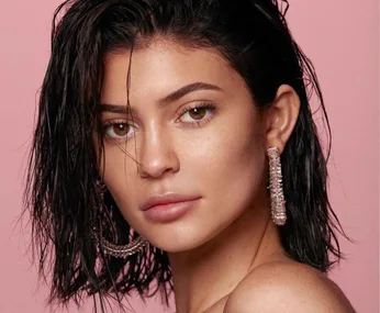 Portrait of a woman with wet hair, wearing large dangling earrings, against a pink background.