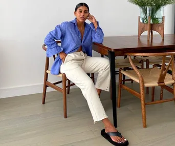 Person in a blue shirt, white pants, and black flip-flops sits on a wooden chair next to a table with green plants.