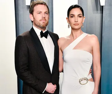 A couple dressed elegantly, the man in a tuxedo and the woman in a white one-shoulder dress, posing at an event.