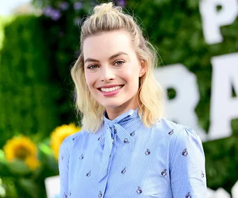 Smiling person with blonde hair in a blue blouse, outdoors with blurred greenery and sunflowers in the background.