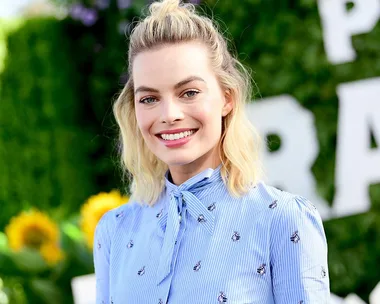 Smiling person with blonde hair in a blue blouse, outdoors with blurred greenery and sunflowers in the background.