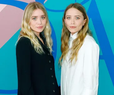 Two women with long hair, one in a black coat and the other in a white shirt, stand together against a colorful backdrop.