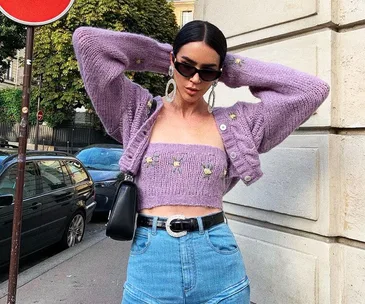 Woman in purple knit crop top and cardigan with sunglasses, standing on a city street.