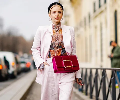 Woman in light pink suit with paisley shirt, holding a vibrant pink handbag, walking on a city street.