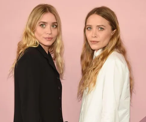 Two women with long blonde hair stand side by side against a pink background, one in black, the other in white.