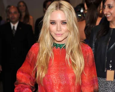 Blonde woman in a red dress, smiling at a formal event with people in the background.