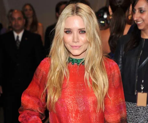 Blonde woman in a red dress, smiling at a formal event with people in the background.