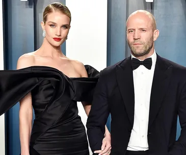 A woman in a black gown and man in a tuxedo pose together at a formal event, holding hands.