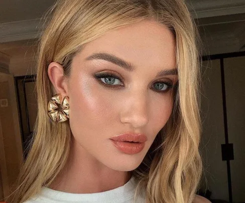 Blonde woman with smoky eye makeup and nude lips, wearing large geometric earrings, gazing directly at the camera.