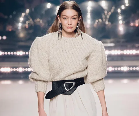 A model walks the runway in an oversized cream sweater with bold, geometric earrings and a wide black belt.