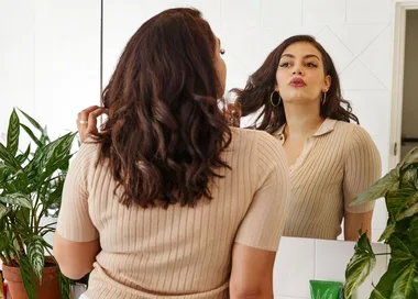 A woman with curly hair looks in a mirror; she's wearing a beige top, surrounded by green plants in a bright room.