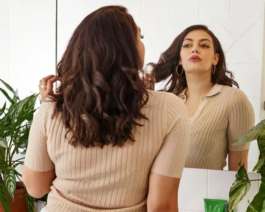 A woman with curly hair looks in a mirror; she's wearing a beige top, surrounded by green plants in a bright room.