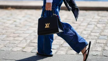 A guest wears a black and gray LV monogram print pattern silk scarf from Louis Vuitton, navy blue large cargo pants, a white shirt, a black shiny leather handbag with a LV gold buckle from Louis Vuitton, black flip flop, a silver and diamonds ring, outside OperaSport, during Copenhagen Fashion Week Spring/Summer 2023, on August 10, 2022 in Copenhagen, Denmark.