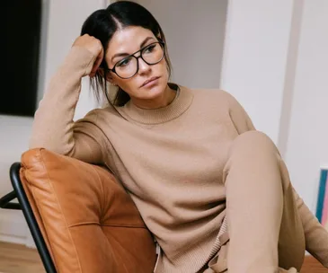 Woman in glasses wearing a beige sweater and pants, sitting on a tan leather chair, looking thoughtful.