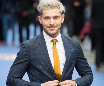 Man in a blue plaid suit and yellow tie, standing on a blue carpet background, smiling at a public event.