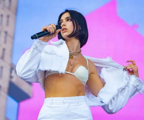 Pop singer performs on stage, wearing a white outfit and holding a microphone against a colorful background.