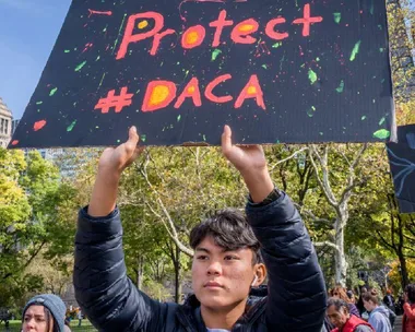 Young man holds "Protect #DACA" sign at outdoor rally, surrounded by trees and people.