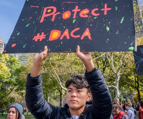 Young man holds "Protect #DACA" sign at outdoor rally, surrounded by trees and people.