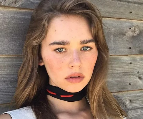 Woman with long hair and freckles, wearing a choker, stands against a wooden wall.