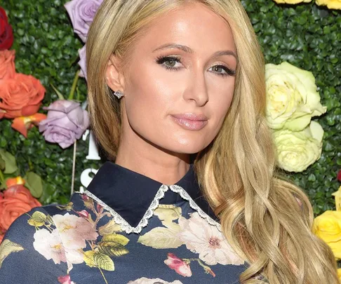 A person with long blond hair and floral dress poses in front of a vibrant floral backdrop.