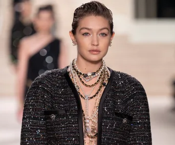 A model wearing a glittering black jacket and layered necklaces walks on a runway, with blurred figures in the background.