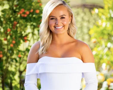 A woman with blonde hair wearing a white off-shoulder dress smiles in a garden with orange trees.