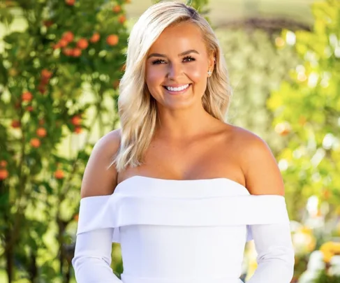 A woman with blonde hair wearing a white off-shoulder dress smiles in a garden with orange trees.