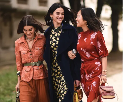 Three women walking together, smiling, in stylish colorful outfits, each holding handbags.