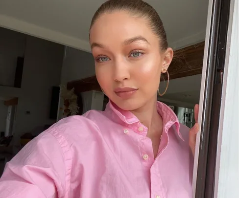 Person in a pink shirt takes a selfie indoors, with hair tied back and hoop earrings.