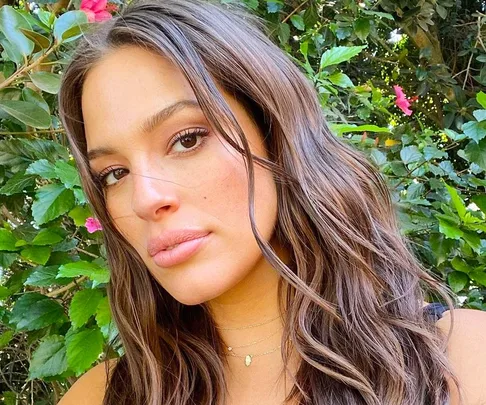 A woman with long wavy hair poses outdoors in front of leafy greenery.