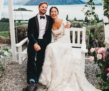 Happy couple in wedding attire seated on a swing, surrounded by flowers, with a scenic lake and mountain backdrop.