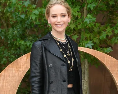 A person smiling, wearing a black leather coat and layered necklaces, with a leafy background.