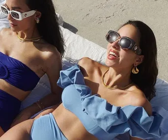 Two women in sunglasses and stylish bikinis relax on a beach towel under the sun.