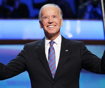 Smiling man in a suit and tie with outstretched arms, standing on a brightly lit stage.