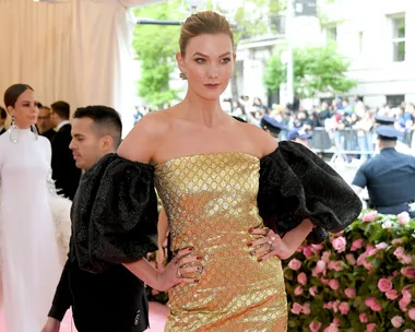 Woman in a gold dress with black sleeves poses confidently on a red carpet, surrounded by attendees and lush decorations.