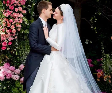 A couple embracing at their wedding; bride in white gown and veil, groom in black suit, surrounded by pink flowers.