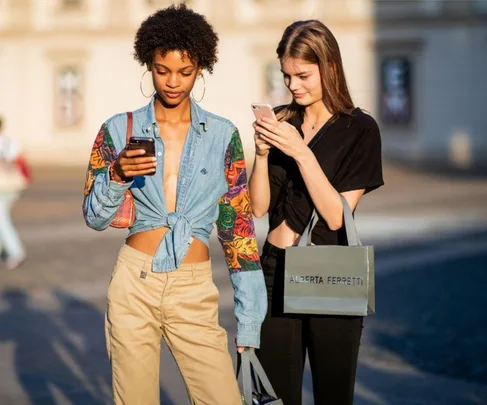 Two fashionable women walking outside, checking their phones, holding shopping bags.