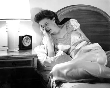 Woman in a nightgown sitting on bed, appearing distressed, with a lamp and clock on the bedside table.