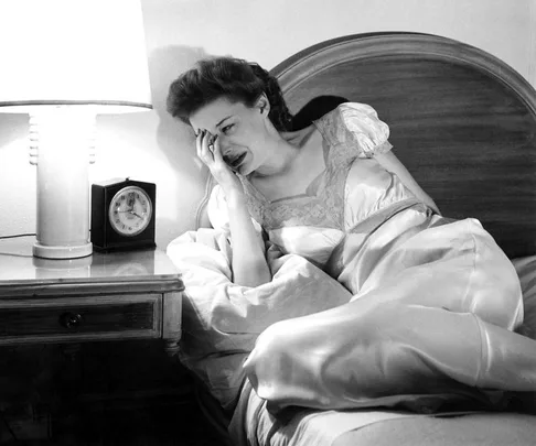 Woman in a nightgown sitting on bed, appearing distressed, with a lamp and clock on the bedside table.