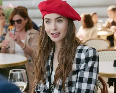 Emily in Paris character wearing a red beret and checkered blazer, seated at an outdoor cafe.