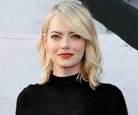 A person with short, wavy blond hair and a black top smiles at the camera against a neutral background.