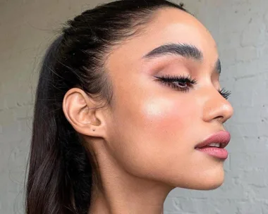 A woman with sleek eyeliner and natural makeup, profile view, hair in a ponytail, against a light brick wall.