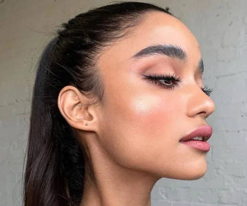 A woman with sleek eyeliner and natural makeup, profile view, hair in a ponytail, against a light brick wall.