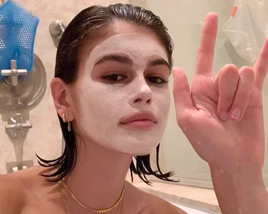 Person with a white face mask in a bathroom, making a hand gesture, with wet hair and gold jewelry.