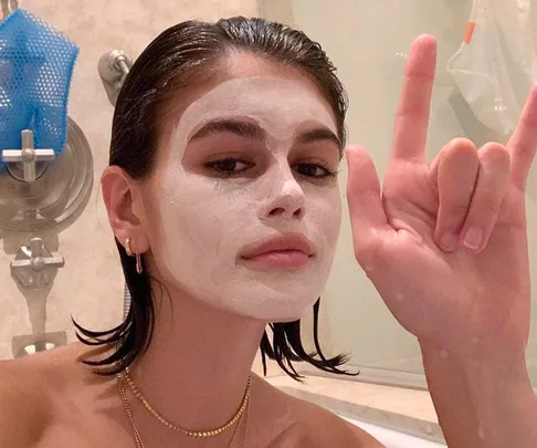 Person with a white face mask in a bathroom, making a hand gesture, with wet hair and gold jewelry.