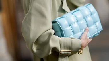 Person holding a light blue woven clutch bag, wearing a beige coat with a gold bracelet.